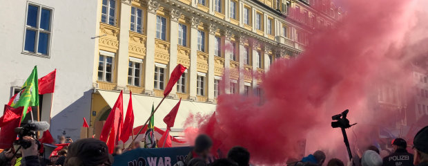 Bild von einer Versammlung im Rahmen der Sicherheitskonferenz 2019, Banner werden von Demonstranten getragen, es steigt roter Rauch aus der Mitte auf