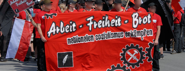 Menschen mit schwarz weiß roten Flaggen und einem Transparent mit der Auschrift: "Arbeit Freiheit Brot - nationalen  Sozialimus erkämpfen"