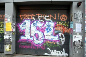 Graffiti Zahl 161 weiß mit Umriss in pinker Farbe auf schwarzem Garagentor mit weiteren orange farbenen Schriftzügen