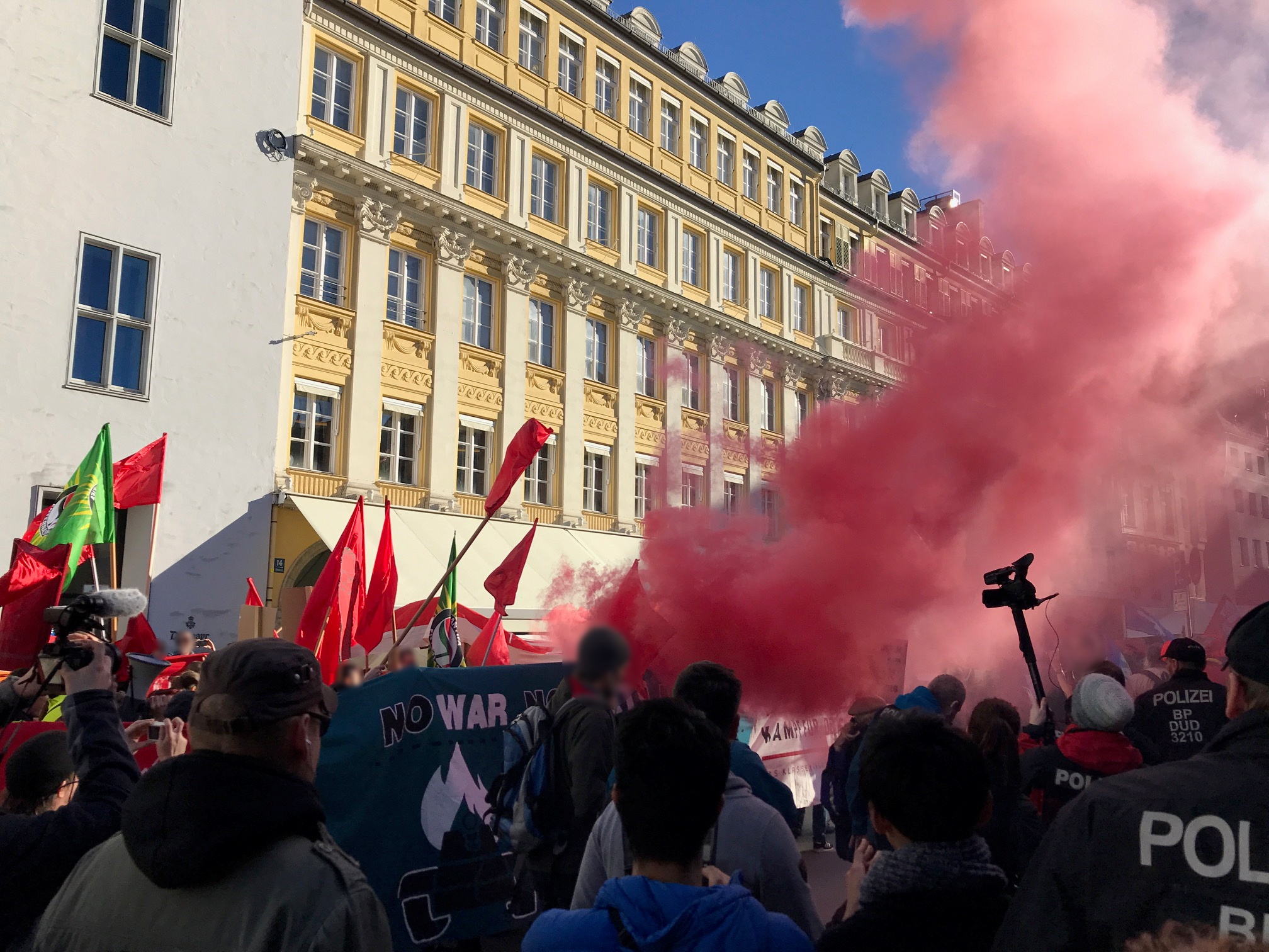 Bild von demonstrierenden Linksextremisten bei der Münchner Sicherheitskonferenz 2019 mit Fahnen und rotem Rauch vor einer Häuserfassade