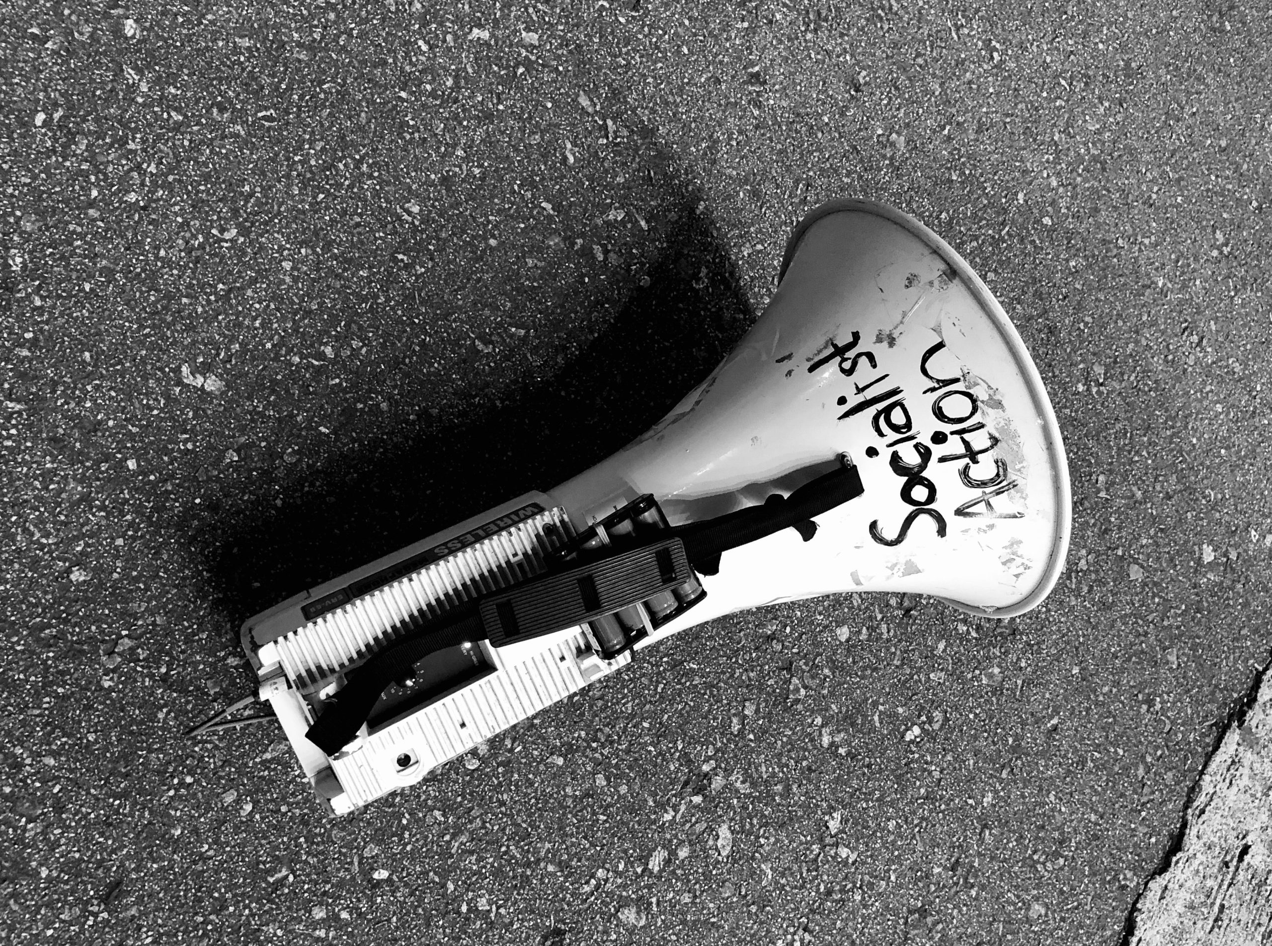 Schwarz-weiß Bild eines auf der Straße liegenden Megafons mit aufgemaltem Spruch auf dem Trichter: Socialist Action
