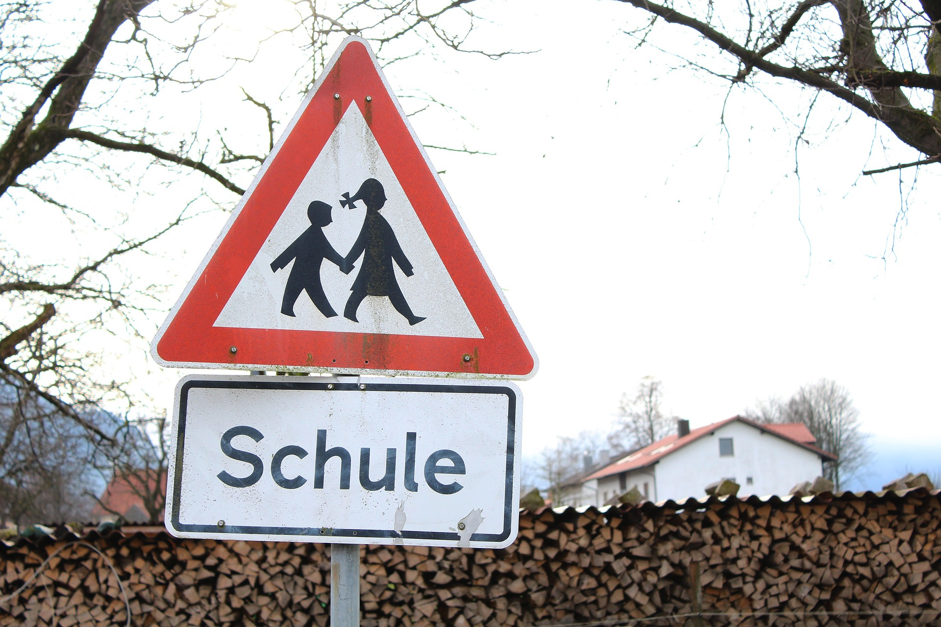 Dreieckiges Verkehrsschild mit rotem Rand mit symbolisierter erwachsener weiblicher Person mit Kind an der Hand in schwarzer Farbe mit unten angebrachtem weißem Schild mit dem Schriftzug Schule. Im Hintergrund Holzhaufenwand mit Häusern dahinter und Bäumen ohne Blätter in kalter Jahreszeit.