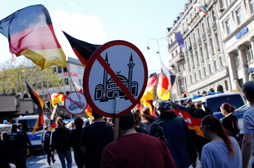 Symbolbild Islamfeindlichkeit. Ein Foto von einer Kundgebung, bei der Demonstranten mit Fahnen und Schildern eine Straße entlang marschieren. Ein Schild zeigt ein durchgestrichenes Moscheesymbol. Im Hintergrund sind Polizeifahrzeuge und weitere Demonstranten zu sehen.