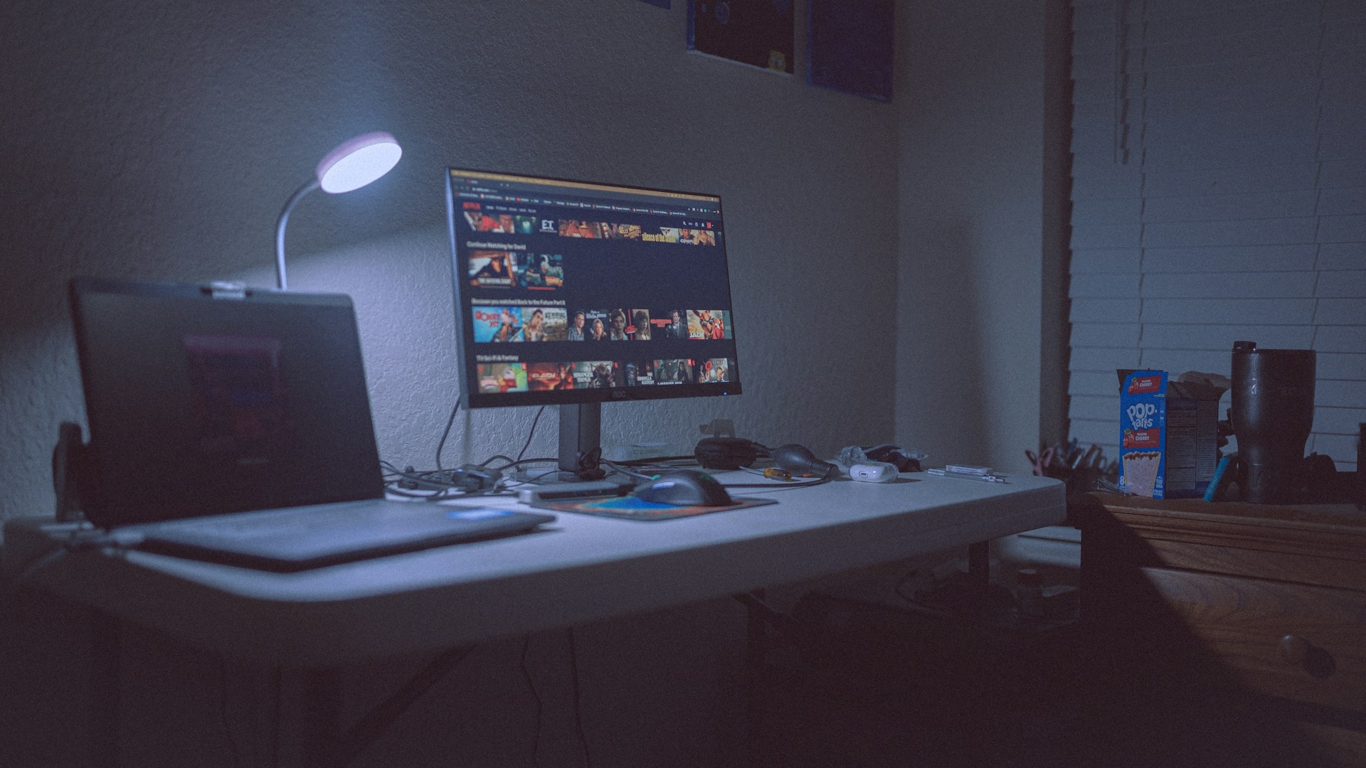 Ein Laptop mit angeschlossenem externen Monitor steht auf einem Schreibtsich in einem abgedunkelten Zimmer, beleuchtet von einer kleiner Stehlampe.