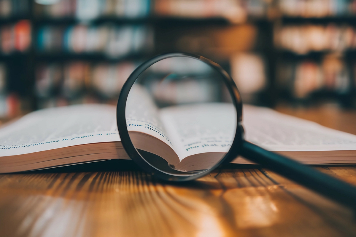 Symbolbild Lupe, Informationen. Eine Lupe liegt vor einem aufgeschlagenem Buch auf einem Holztisch. Im Hintergrund ist verschwommen ein Regal mit Büchnern zu erkennen.