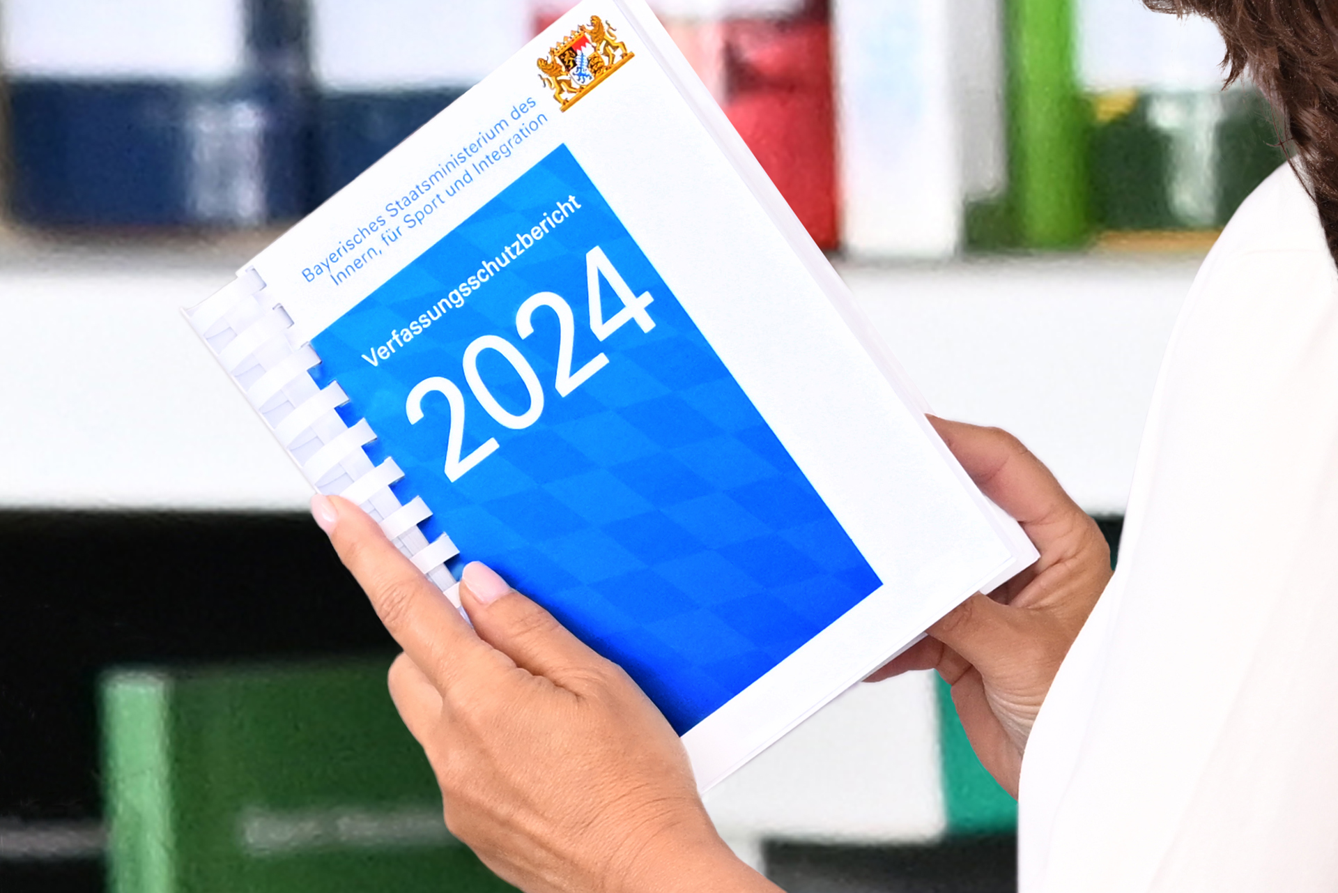 Eine Person hält in ihren Händen ein gedrucktes Exemplar des Bayerischen Verfassungsschutzberichtes vor einem Bücherregal.