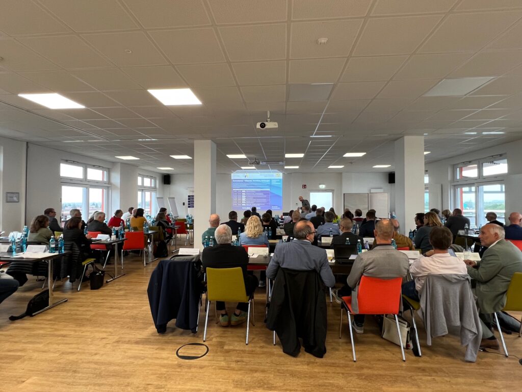 Menschen in Aula an Tischen sitzend von hinten betrachet mit einem Referenten vorne.